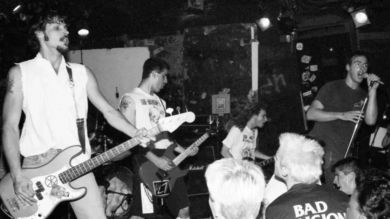 Live-Auftritt einer Punkband in den 80ern. Bassist mit weißem Hemd, Gitarrist und Sänger im Vordergrund.