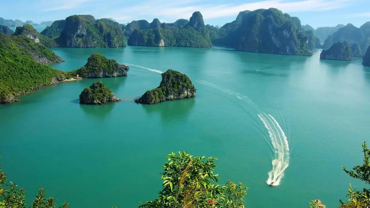Baia di Ha Long, Vietnam: isole calcaree ricoperte di vegetazione emergono da acque turchesi. Un motoscafo lascia una scia bianca. Imperdibile per chi visita il Vietnam cosa vedere.
