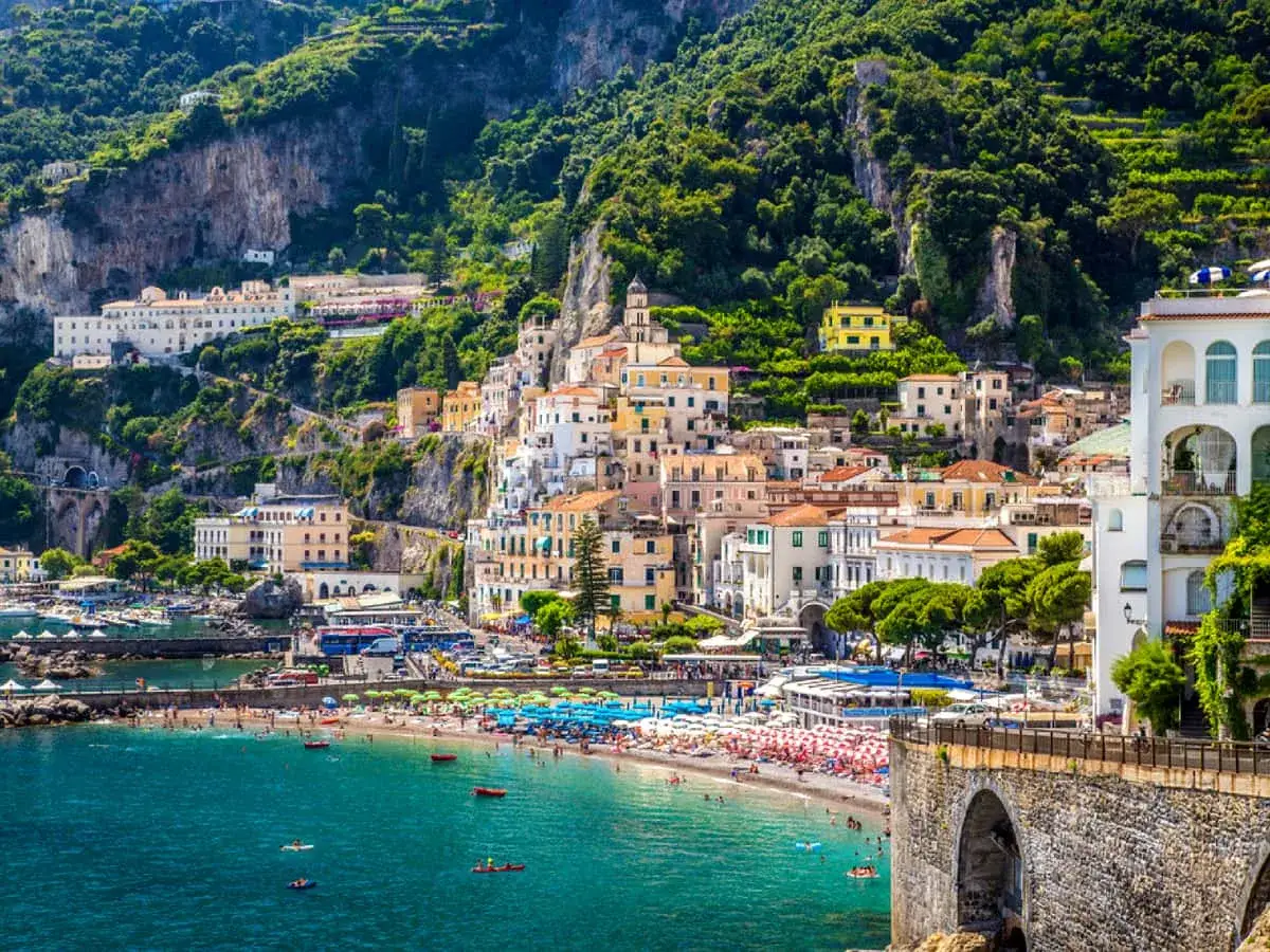 A vibrant beach scene on the Amalfi Coast, with colorful buildings cascading down a lush green cliffside. Tourists enjoy the sun and sea, suggesting the Amalfi Coast is safe for a relaxing vacation.