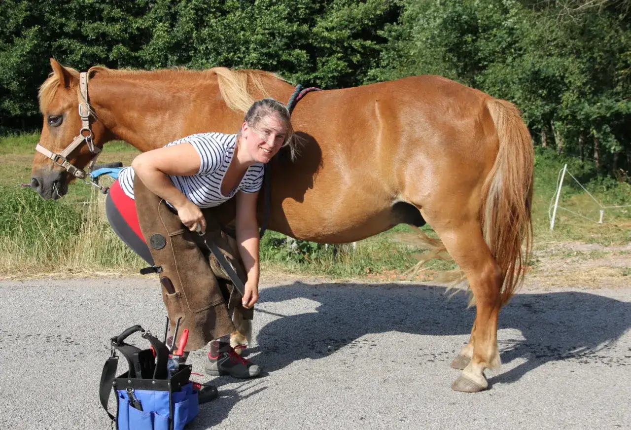 Une maréchale-ferrante travaille sur le pied de cheval sans sabot. Elle sourit, entourée de ses outils.