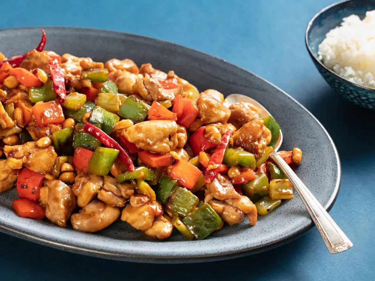 Ein Teller mit w&uuml;rzigem Kung Pao H&auml;hnchen, garniert mit Erdn&uuml;ssen, Paprika und getrockneten Chilis. Daneben eine Sch&uuml;ssel Reis.
