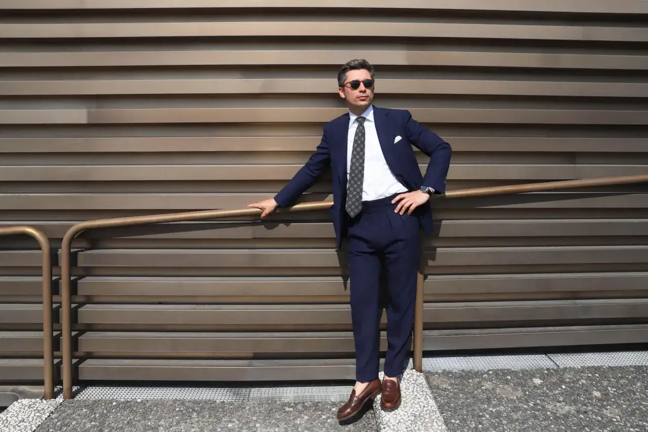 Un homme élégant porte un costume bleu marine, une chemise blanche et une cravate à motifs. Ses chaussures marron complètent parfaitement sa tenue.