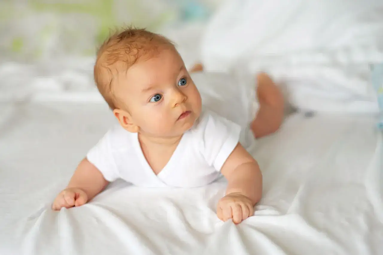 Niebieskooki, 10 tygodniowe dziecko w białej koszulce leży na brzuchu na białej pościeli, zaciekawiony otoczeniem.