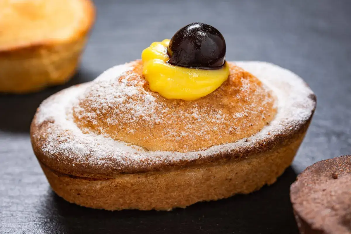 Un delizioso pasticciotto leccese cosparso di zucchero a velo, con crema gialla e una ciliegia scura in cima.
