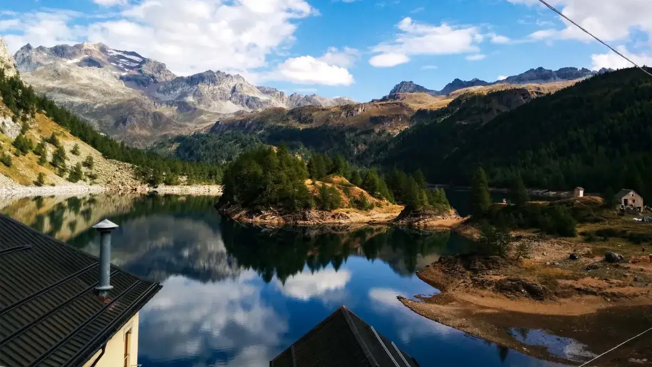 Alpe Devero: lago alpino con montagne maestose, isolotto verdeggiante e riflessi perfetti nel blu intenso dell'acqua.