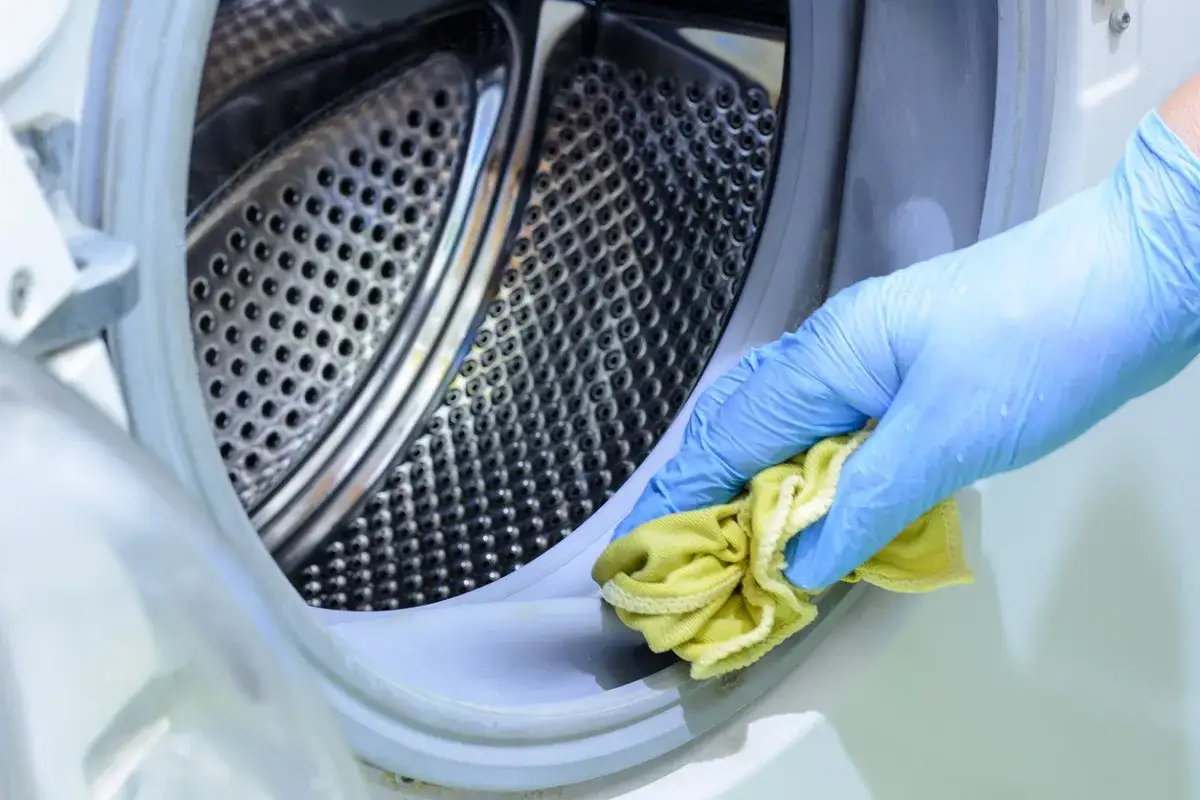 Mano con guante azul limpiando la goma de la lavadora con un paño amarillo. Aprende como quitar el mal olor de la lavadora.