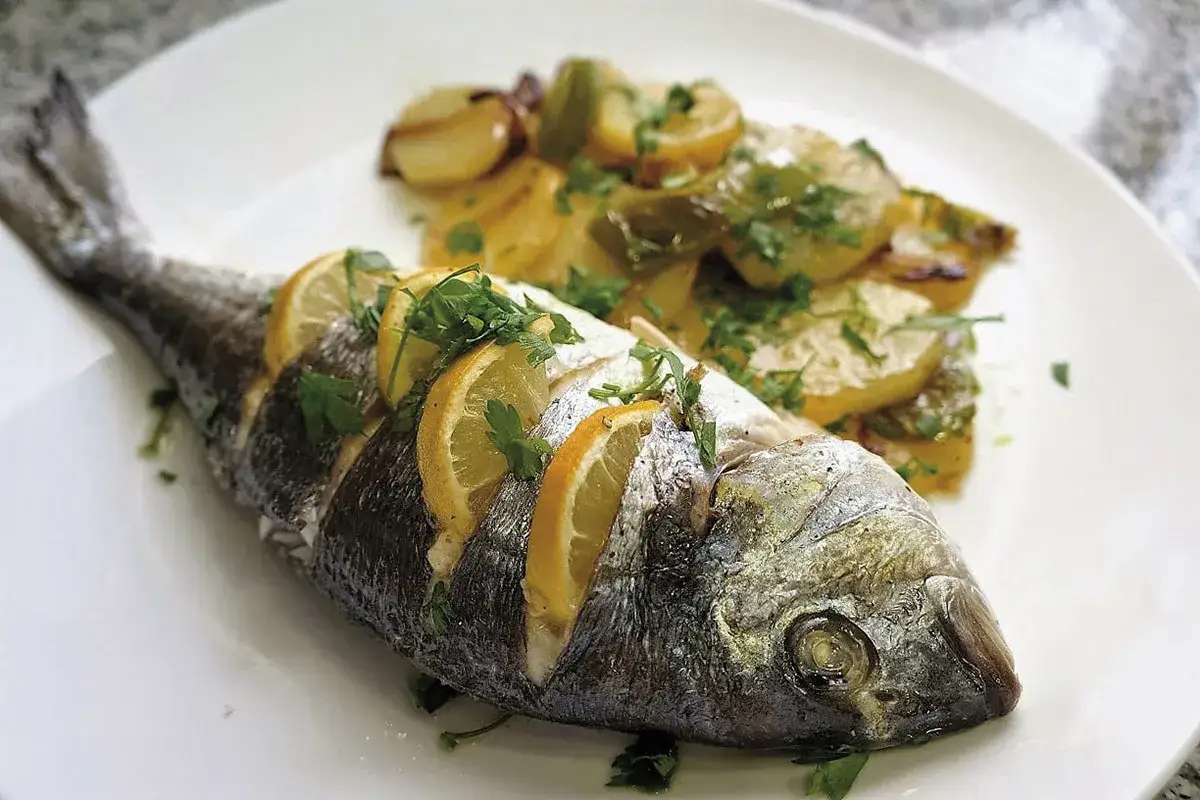 Pescado al horno entero, adornado con rodajas de limón y perejil, acompañado de patatas asadas.