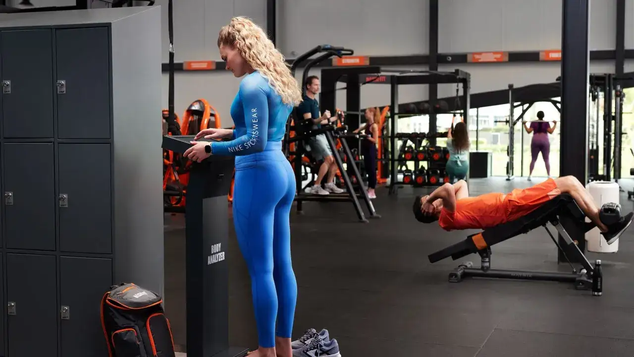 Femme en tenue de sport bleue sur un appareil de mesure corporelle. D'autres personnes s'entra&icirc;nent en arri&egrave;re-plan. Je mange peu, je fais du sport et je grossis, quel paradoxe !