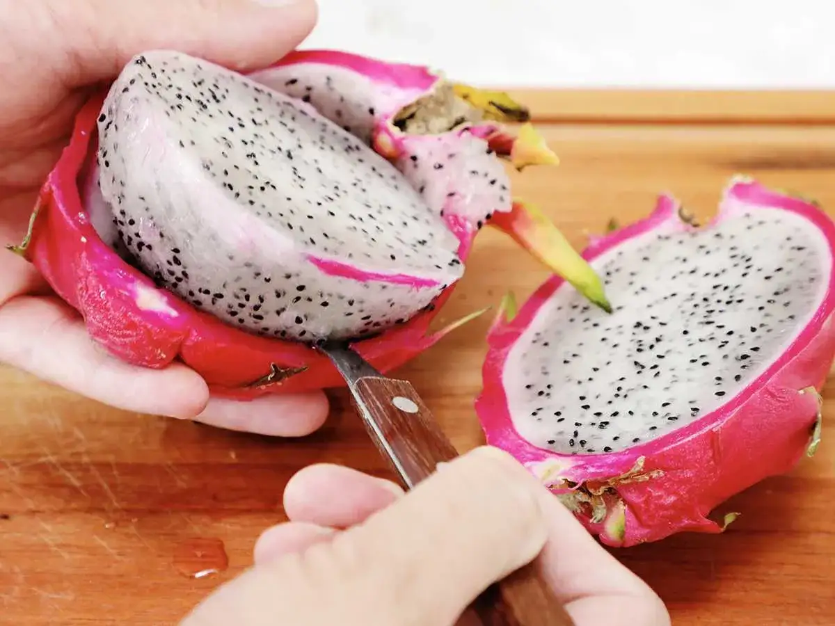 Jak jeść pitahaya? Owoc przekrojony na pół, ręka z nożem delikatnie oddziela miąższ biały z czarnymi pestkami od różowej skórki.