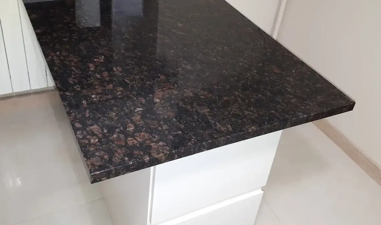 Elegancki blat granitowy 2 czy 3 cm o ciemnobrązowym, nakrapianym wzorze, idealnie komponuje się z białymi frontami szafek.