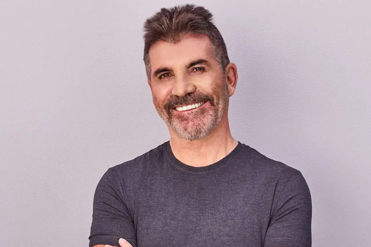 Uśmiechnięty Simon Cowell w szarej koszulce. Plotki o tym, że Simon Cowell nie żyje, są fałszywe.
