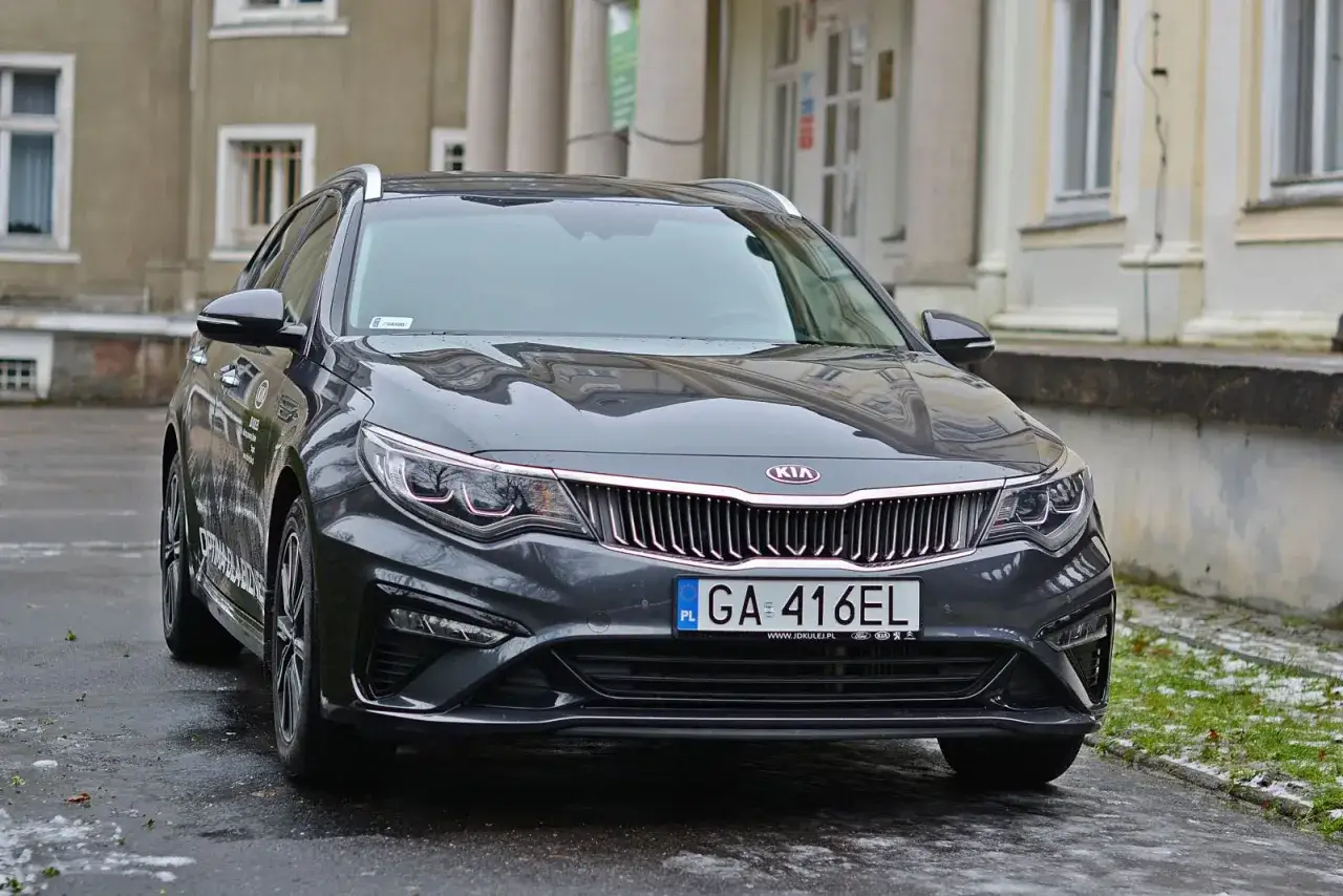 Szary Kia Optima kombi przed budynkiem. Sprawdź kia optima dane techniczne, by poznać jego możliwości.
