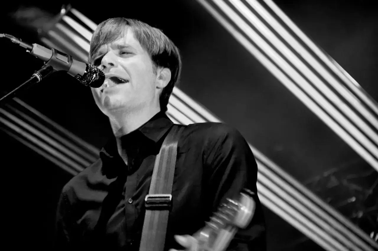 Ben Gibbard chante passionnément dans un micro, sa guitare à la main, sous des lumières de scène.
