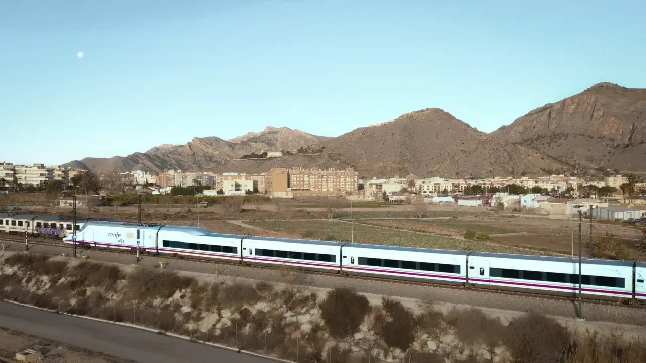 Tren AVE Renfe, un moderno tren blanco con detalles morados, atraviesa un paisaje rural con monta&ntilde;as y edificios al fondo, rumbo a Madrid desde Orihuela.