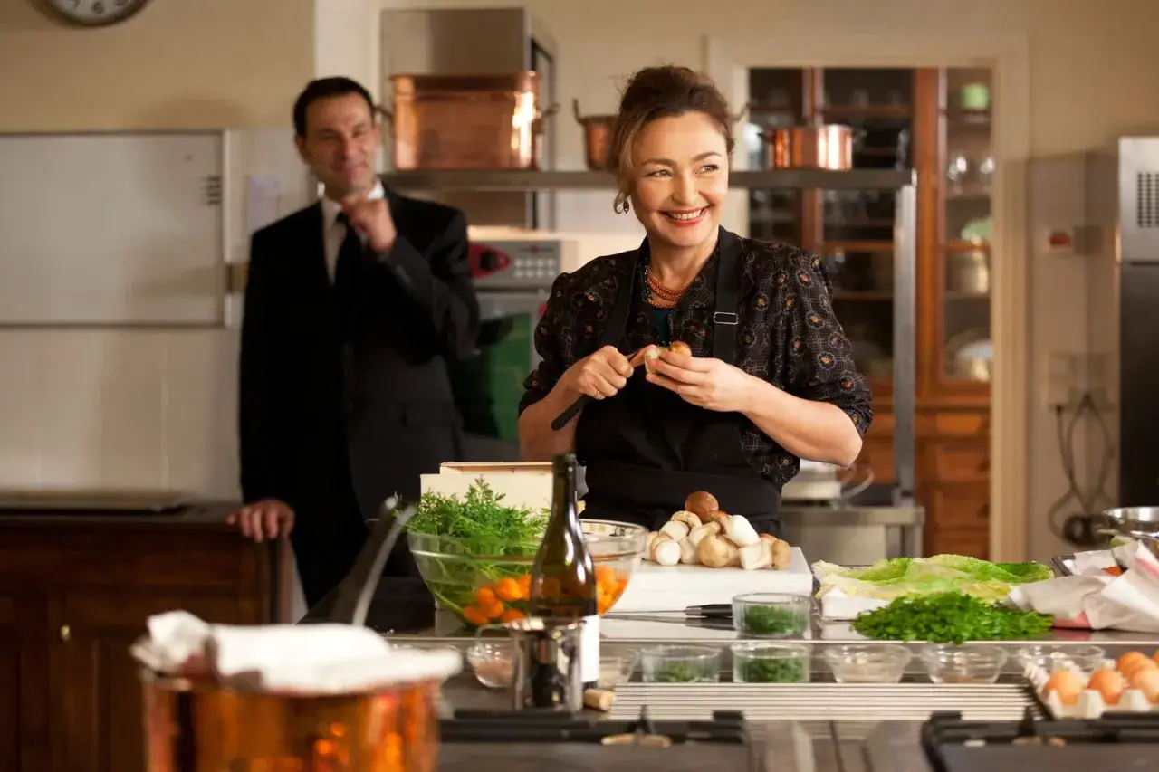 Una mujer sonr&iacute;e mientras prepara ingredientes en una cocina, quiz&aacute;s inspirada por el estilo de cocina de Robuchon que es.