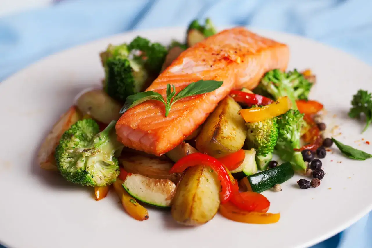Ein saftiges Stück low carb Lachs auf einem Bett aus geröstetem Gemüse wie Brokkoli, Paprika und Zucchini, garniert mit frischen Kräutern.