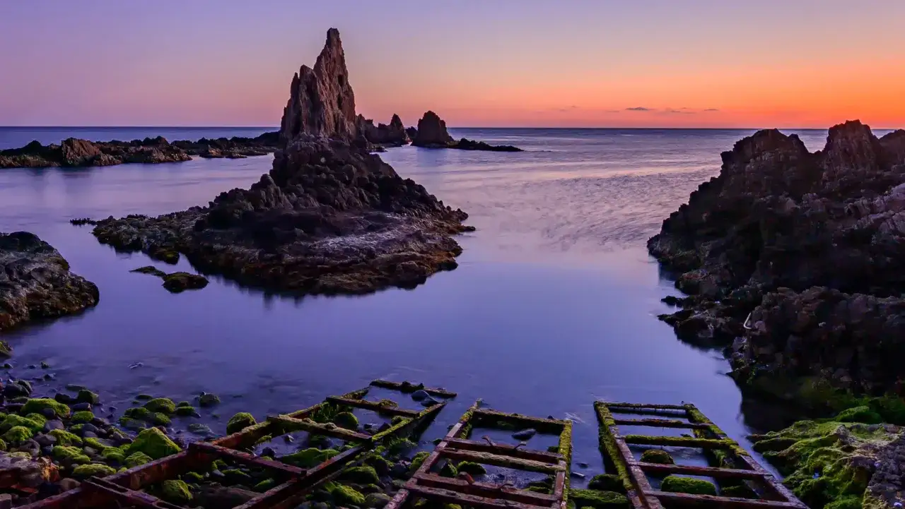 Rocas afiladas emergen del mar sereno al amanecer en la sierra Cabo de Gata. El cielo se tiñe de tonos rosados y púrpuras, reflejándose en el agua tranquila.