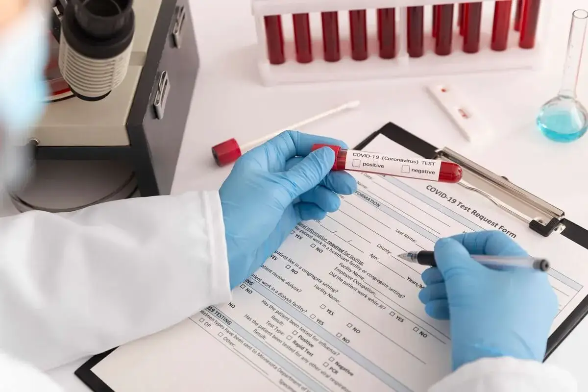 Ręce w niebieskich rękawiczkach trzymają probówkę z krwią do testu COVID-19. W tle mikroskop i probówki. Wypełniany formularz z informacją o podwyższonym MPV.