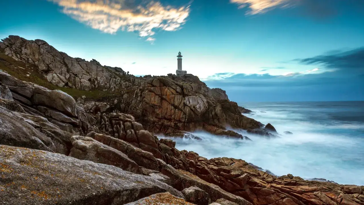 El faro de Punta Nariga se alza sobre rocas escarpadas, con el mar agitado y el cielo crepuscular creando una escena dramática.
