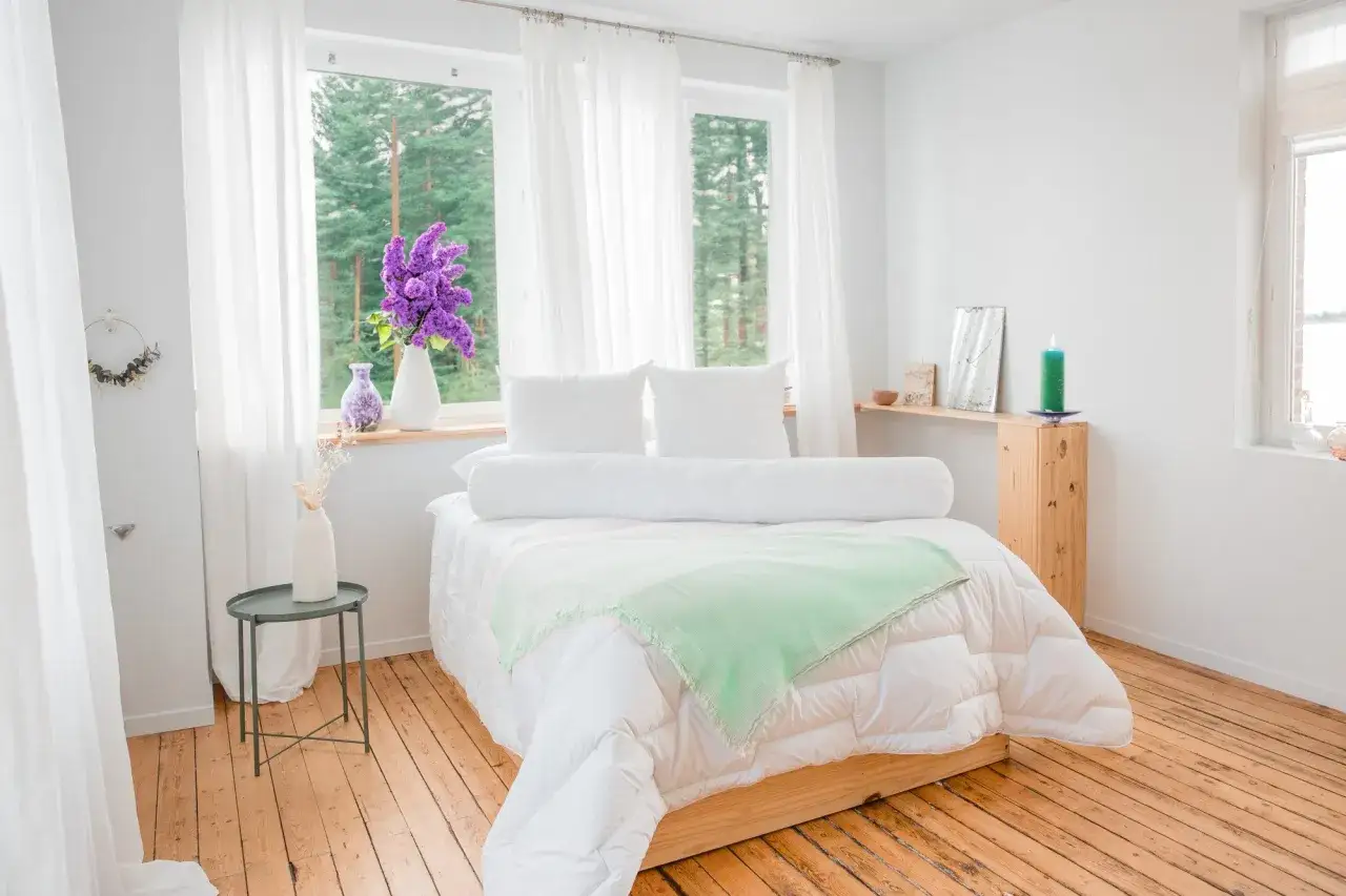 Un lit douillet avec un traversin blanc, des oreillers moelleux et une couverture verte. Des rideaux blancs et des fleurs violettes apportent une touche de fraîcheur.