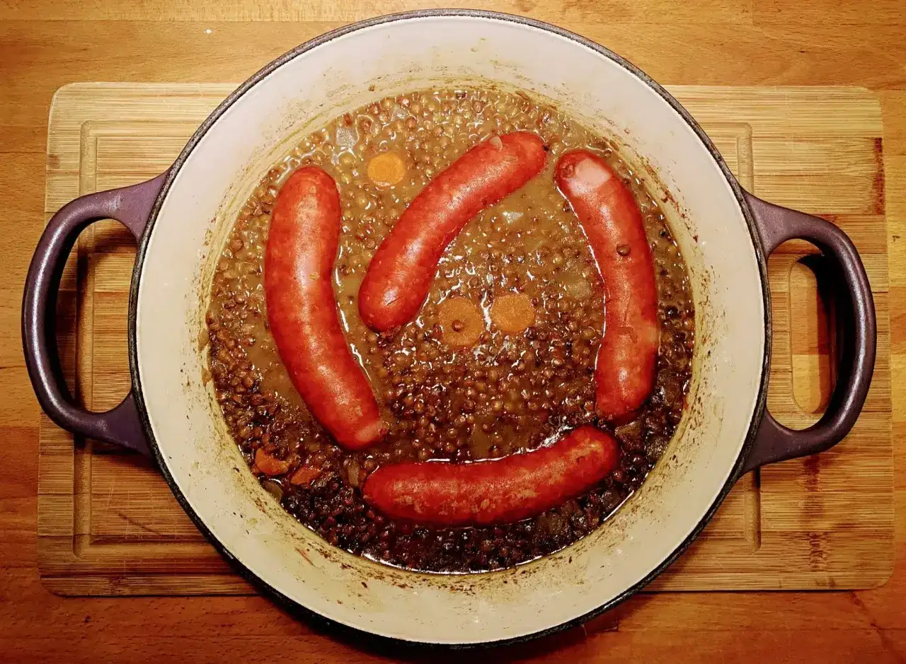 Un plat de saucisse lentille mijote dans une cocotte en fonte, avec des rondelles de carottes. Un repas réconfortant.