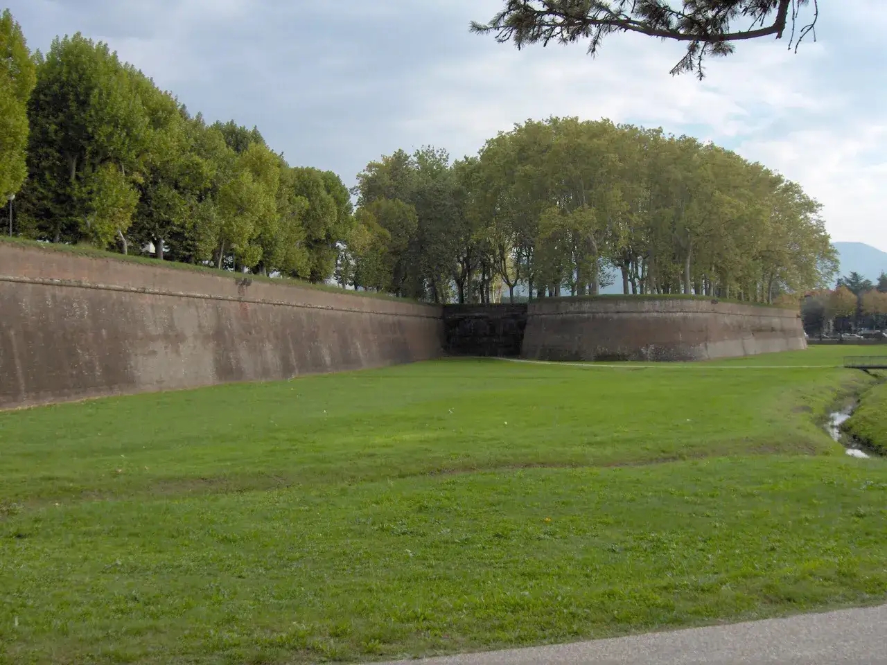 A grand, ancient walled city in Tuscany, its imposing brick ramparts curve around a lush green park, topped by a dense line of trees.