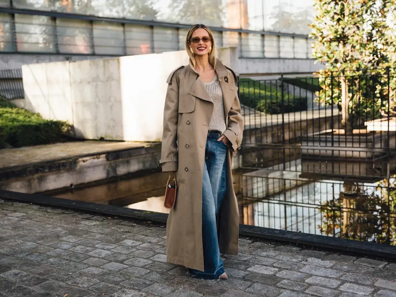 Frau im langen Trenchcoat, der l&auml;ssig zu Jeans und Strickoberteil kombiniert wird. Sonnenbrille und kleine Tasche runden den Look ab.