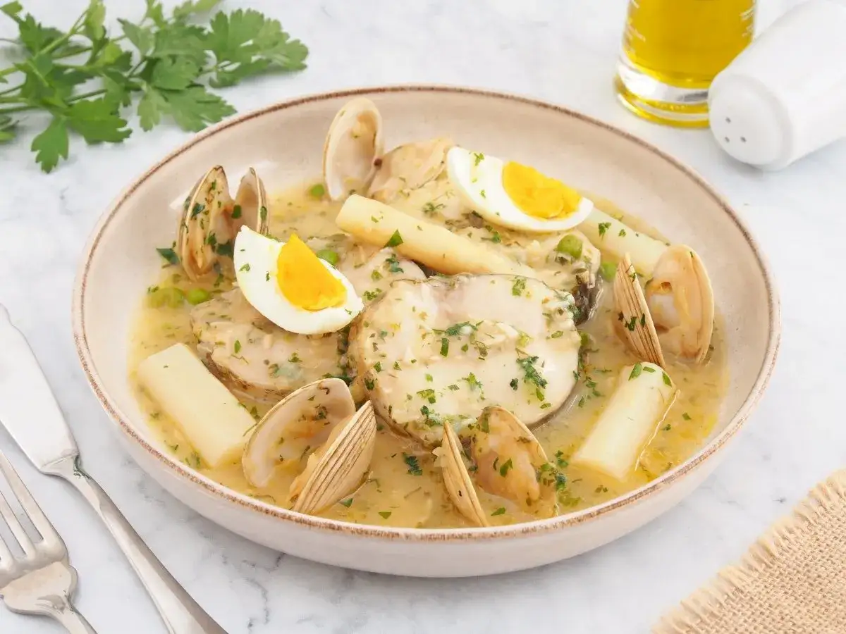 Merluza a la vasca con almejas, esp&aacute;rragos blancos y huevo duro, servida en un plato beige.