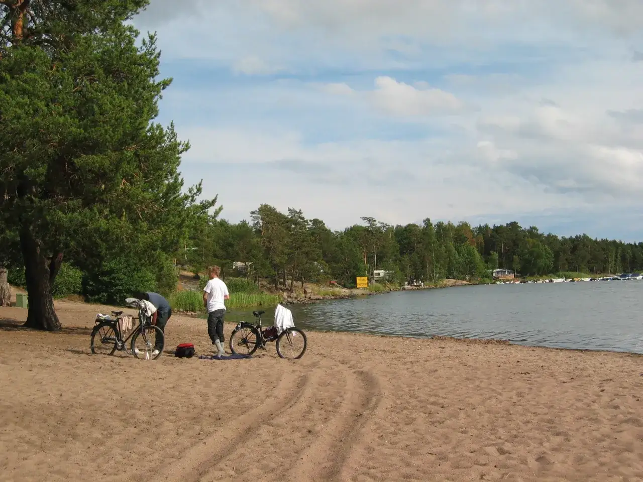 Tv&aring; personer med cyklar p&aring; en sandstrand vid Hallmare Havsbad. Vattnet glittrar och en skogskl&auml;dd kustlinje syns i bakgrunden.