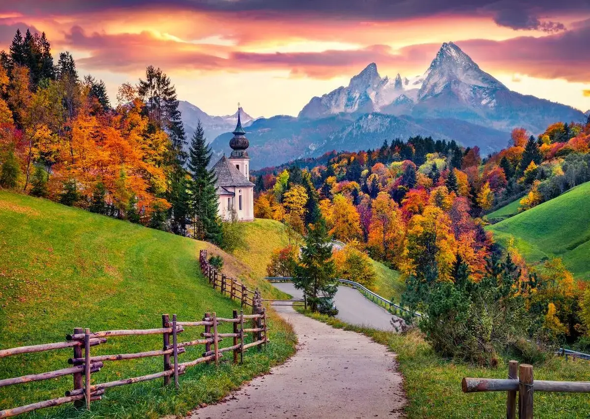 Alpenpanorama im Oktober: Ein Weg führt zu einer Kirche, umgeben von bunten Bäumen und schneebedeckten Gipfeln. Perfekt für Backpacking.