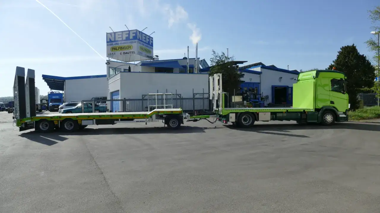 Ein leuchtend gr&uuml;ner Gliederzug steht vor einem Geb&auml;ude. Der LKW mit Auflieger ist bereit f&uuml;r den Transport.