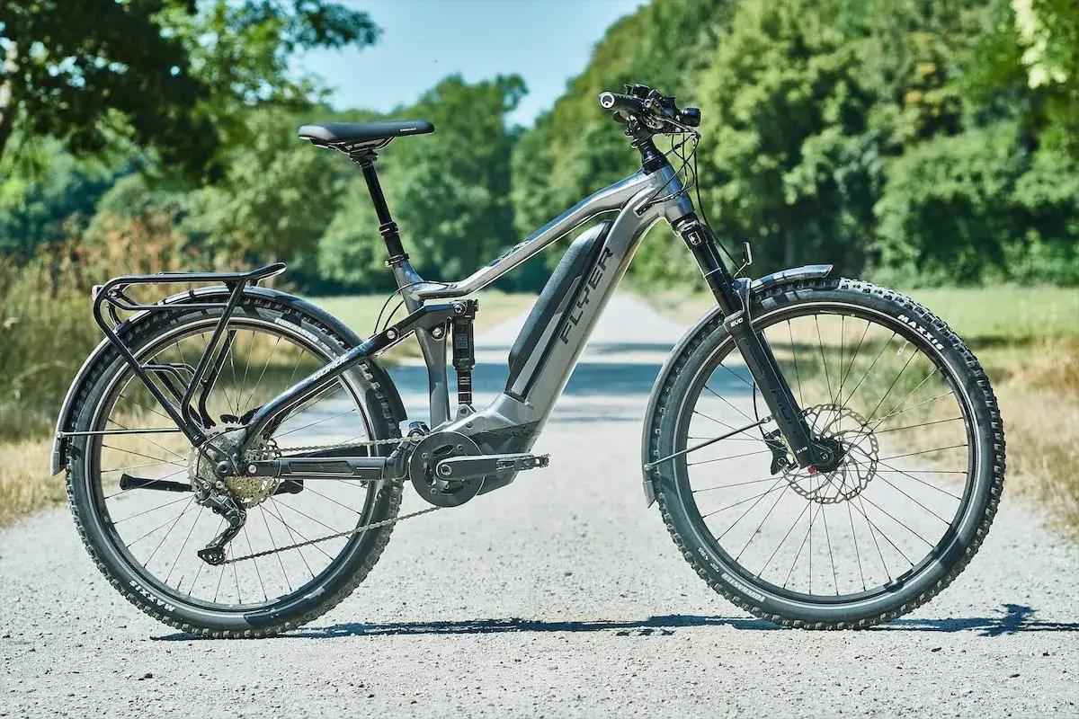 Szary rower elektryczny Flyer na testowej, szutrowej drodze. Pełne zawieszenie i bagażnik sugerują gotowość do długich wypraw.