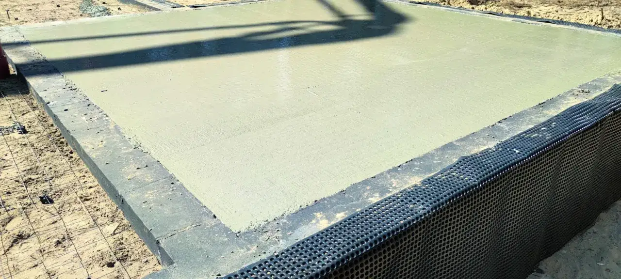 Świeżo wylany beton na fundamentach, zapewniający odpowiednią minimalną grubość chudziaka. Widoczna siatka zbrojeniowa i drenaż.