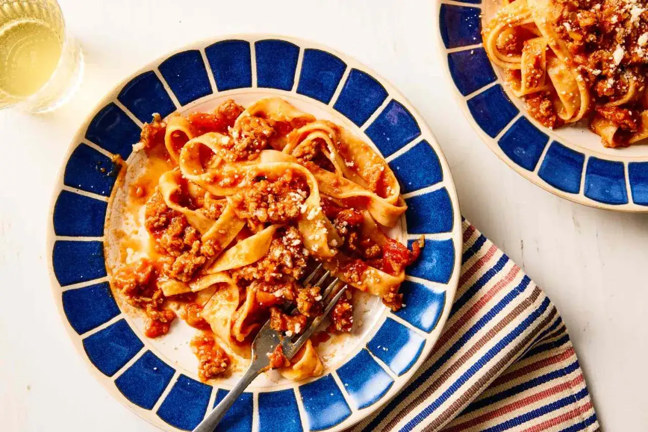Teller mit breiten Bandnudeln und einer herzhaften, **schnellen Bolognese** Sauce, bestreut mit Parmesan. Ein Glas Weißwein steht daneben.