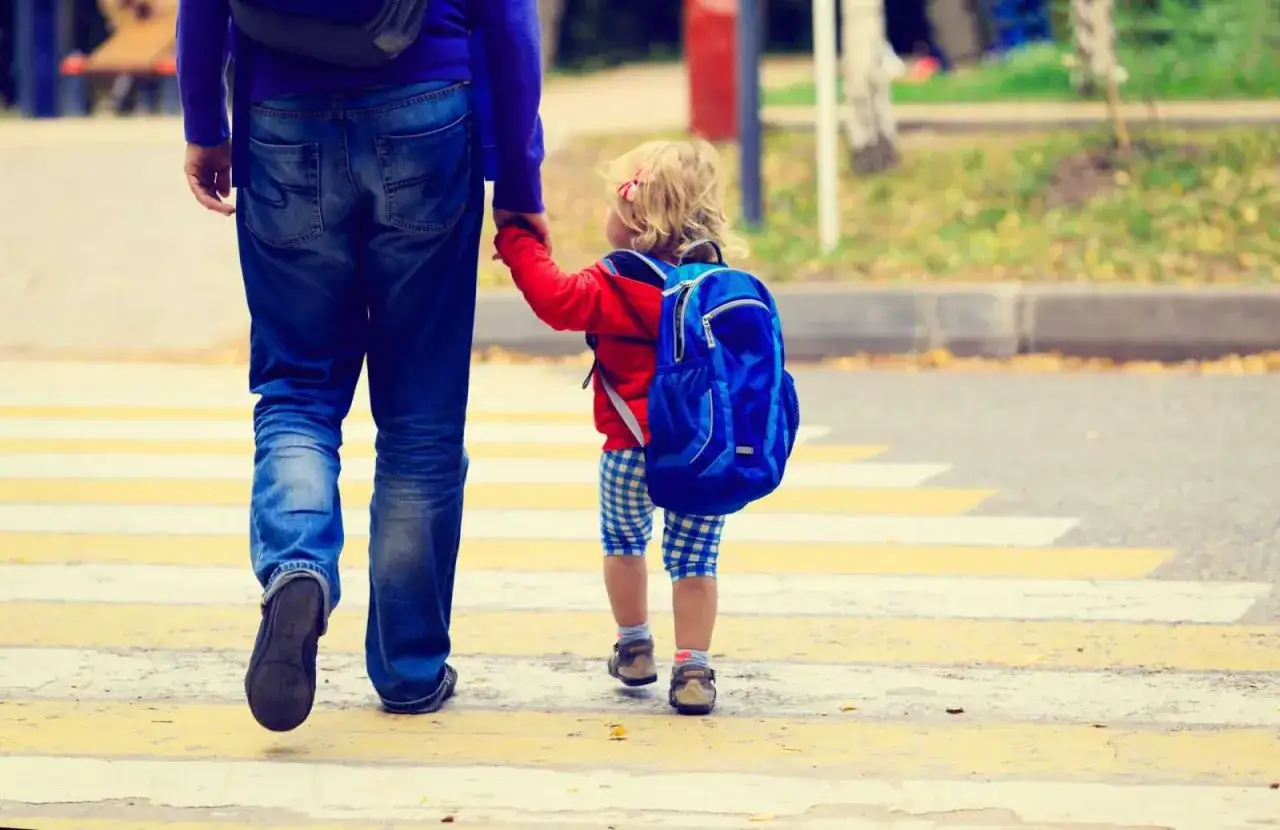 Un padre y su hija cruzan un paso de peatones. La niña, con una mochila azul, inicia su periodo de adaptación escolar.