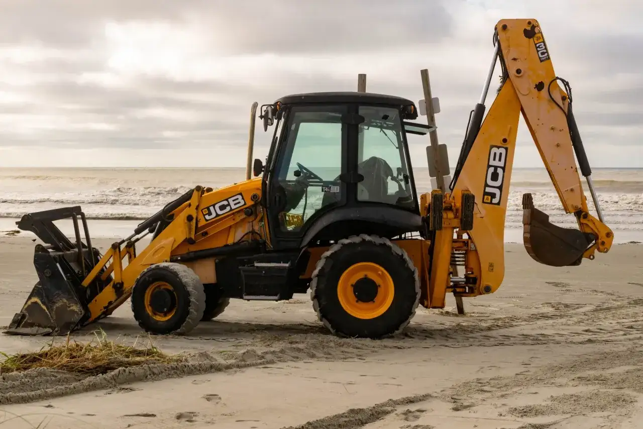 Ż&oacute;łta koparko-ładowarka firmy JCB stoi na piaszczystej plaży, z widokiem na wzburzone morze i zachmurzone niebo.