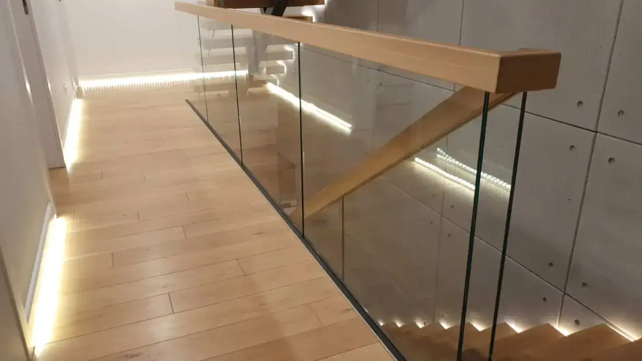 Nowoczesna klatka schodowa w domu z drewnianą poręczą i szklanymi balustradami. Podłoga z jasnego drewna, oświetlenie LED.