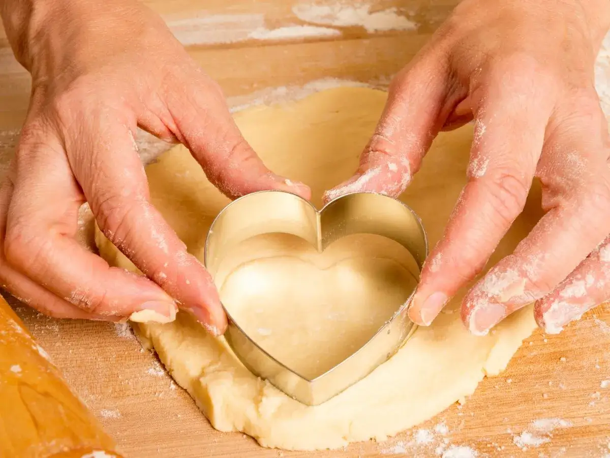 Manos cortan masa de galletas con un cortador en forma de corazón.