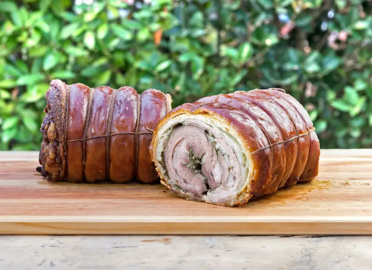 Dwa pieczone rolady z wieprzowiny, czyli porchetta, na desce. W środku widać warstwy mięsa i zi&oacute;ł.