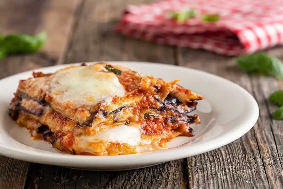 Una fetta di parmigiana di melanzane, un classico delle ricette della nonna, servita su un piatto bianco con basilico fresco.