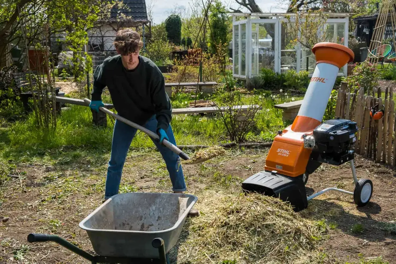 Ein Mann schaufelt Gartenabfälle in einen Häcksler, um Kompost zu zerkleinern. Im Hintergrund ein Gewächshaus und ein Garten.