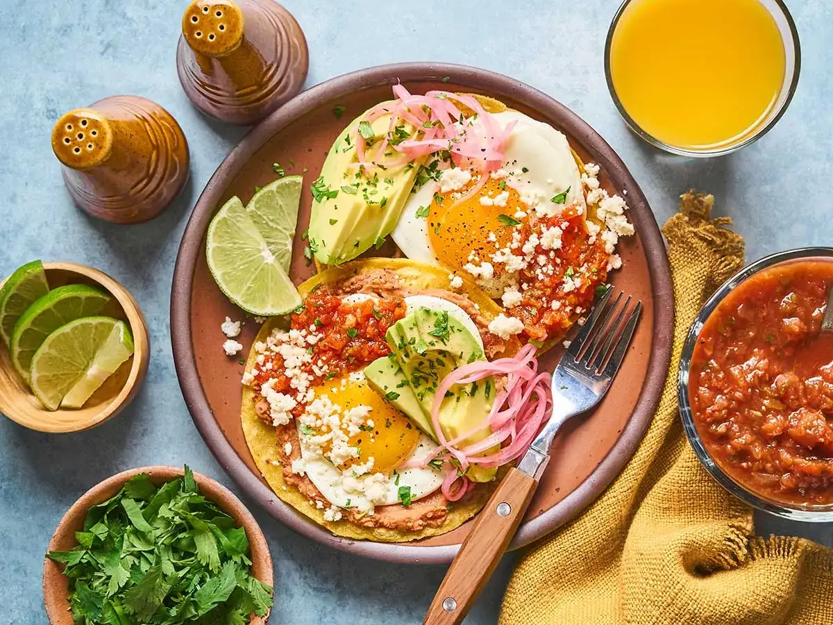 Deux assiettes de huevos rancheros garnies d'avocat, de fromage et de salsa, accompagn&eacute;es de quartiers de lime, de coriandre fra&icirc;che et d'un verre de jus d'orange.