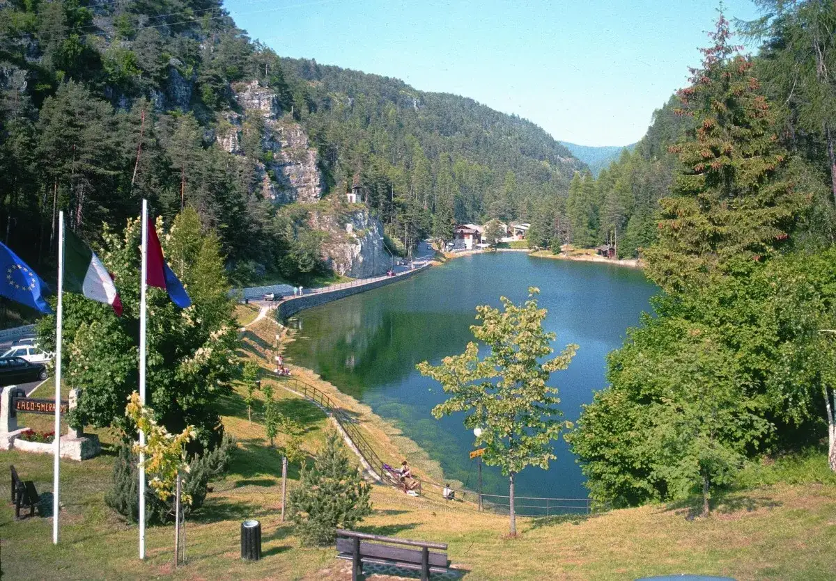 Il **lago smeraldo** incastonato tra montagne boscose, con case e un muretto che lo costeggiano. Bandiere sventolano in primo piano.