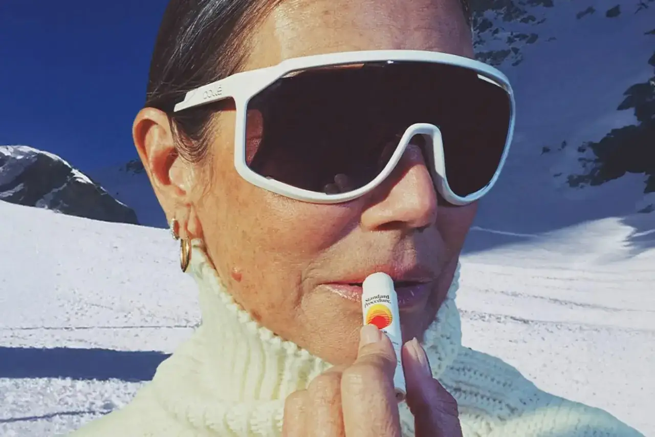 Frau mit Sonnenbrille und Rollkragenpullover trägt Lippenbalsam auf, um ihre Lippen vor der Sonne zu schützen. Ein kleiner Herpesausbruch ist sichtbar.