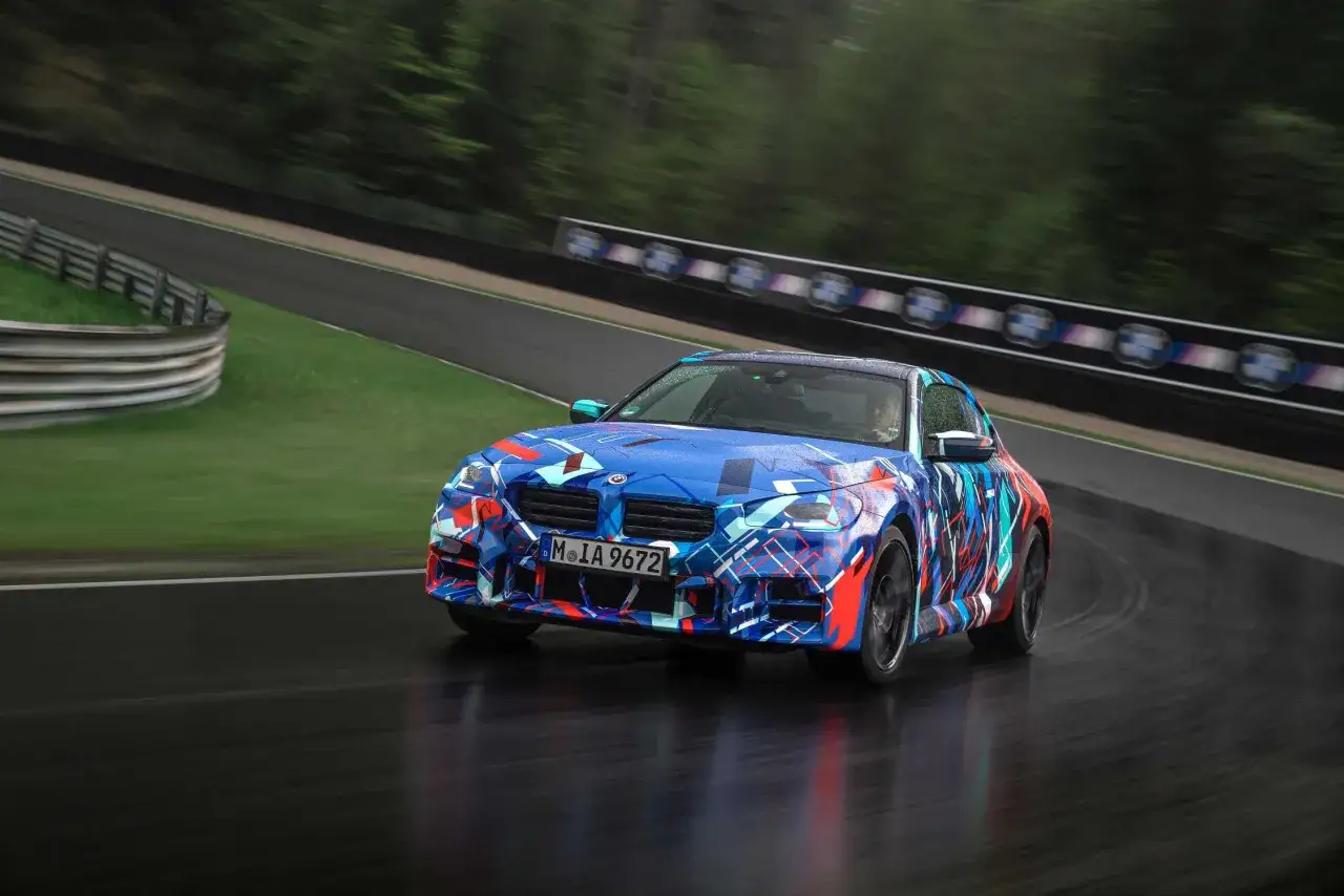 Dynamiczne BMW M2 CS w kamuflażu pędzi po mokrym torze wyścigowym, ukazując swoją sportową naturę.