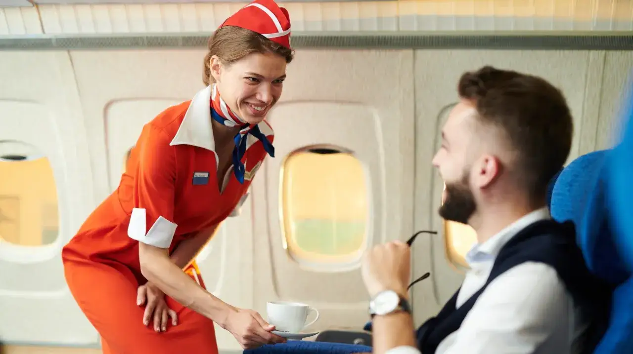 Uśmiechnięta stewardessa w czerwonym mundurku podaje kawę pasażerowi. Marzysz, jak zostać stewardessą? To praca pełna uśmiechu i podróży.