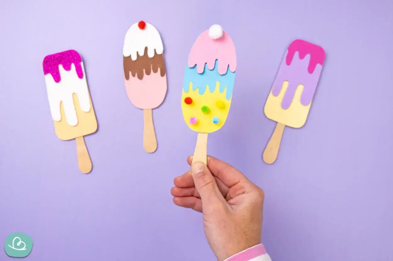 Bunte Eis-Kreationen zum Eis basteln: Vier selbstgemachte Eis am Stiel aus Papier und Pompons auf lila Hintergrund.