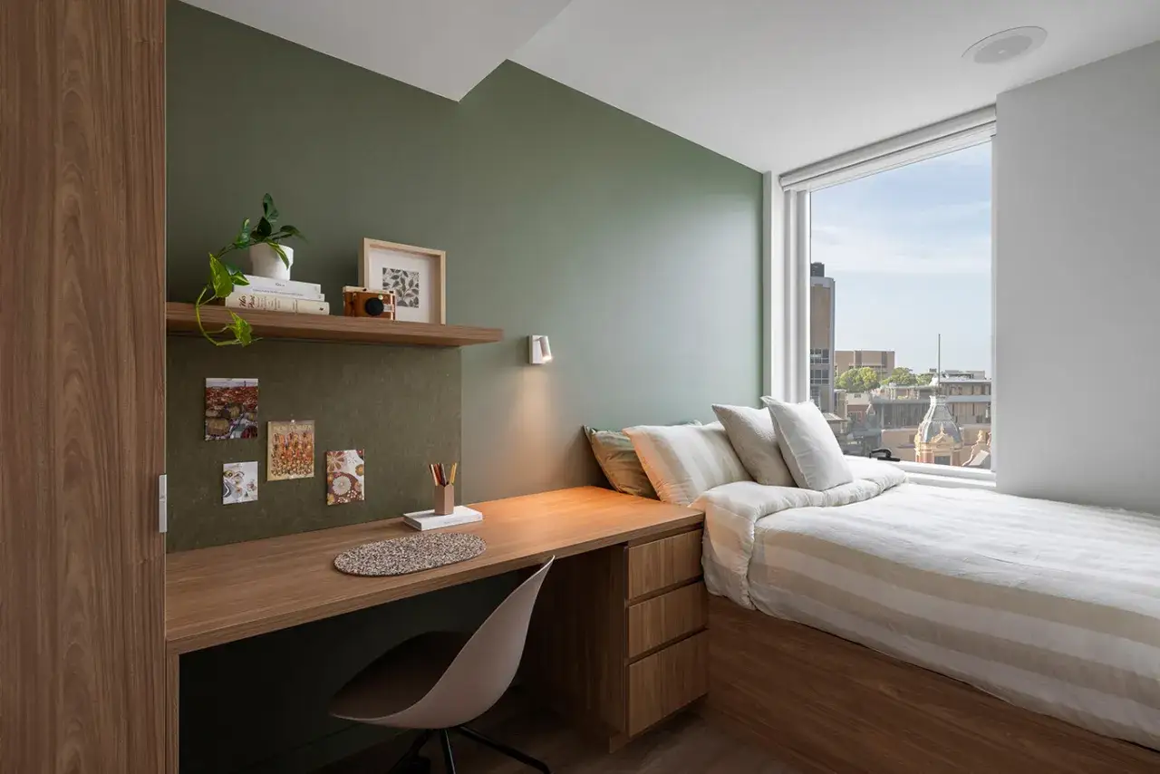 Un dormitorio moderno con escritorio y cama, ideal para vivir en Australia. La vista desde la ventana muestra edificios urbanos.