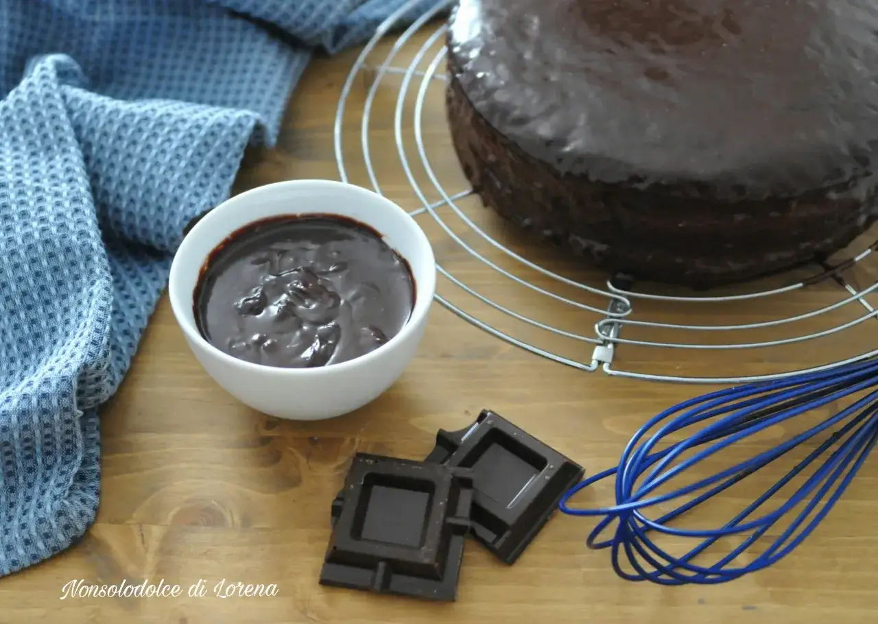 Una torta al cioccolato lucida, una ciotola di ganache cioccolato per copertura, pezzi di cioccolato e una frusta blu su un tavolo di legno.