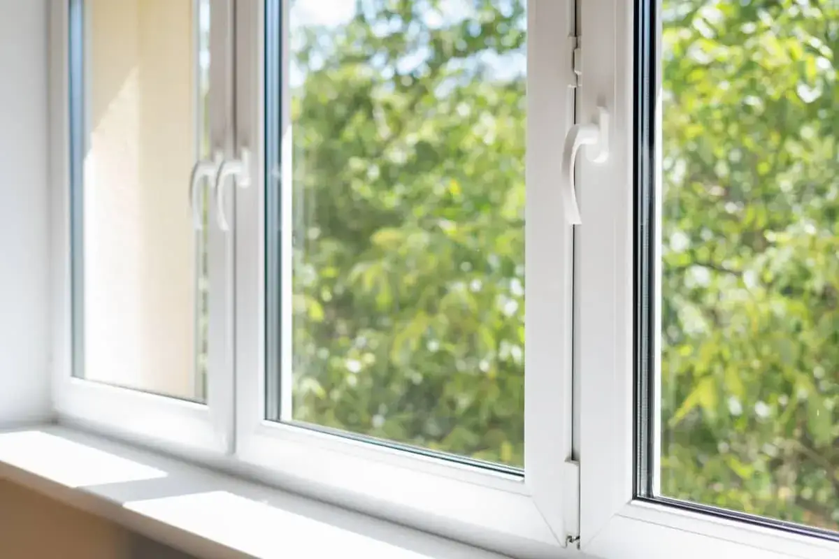 Ventanas sostenibles de PVC blanco, con manillas plateadas, que dejan pasar la luz del sol y muestran un fondo de &aacute;rboles verdes.